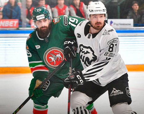 На изображении два хоккеиста в белой и зелёной форме соревнуются за шайбу на льду. На заднем плане видны зрители за стеклянным ограждением